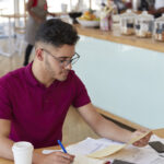 vertical shot of attractive hipster student prepares financial project, rewrites information from document in notepad, sits at desk in cozy restaurant, wears eyewear, poses indoor. paperwork concept
